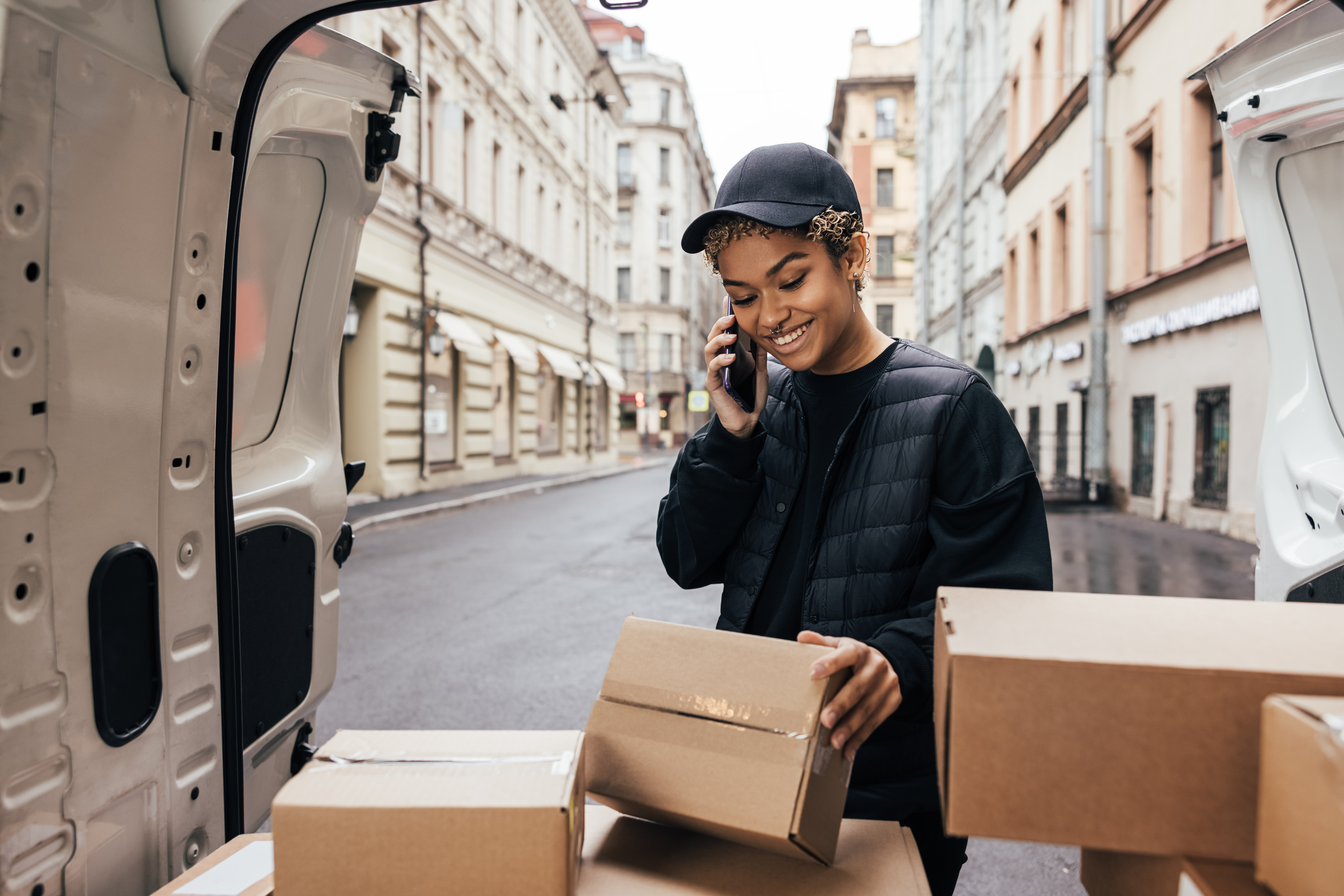 Delivery woman in uniform looking at box and making call to a customer while standing near car trunk